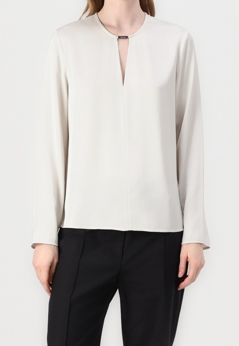 Blouse gris clair à manches longues avec un col rond, présentant une petite ouverture en forme de trou de serrure à l'avant et un tissu lisse légèrement brillant.