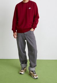 Person som bär en vinröd Nike-tröja, lösa grå jeans med uppvikta benslut och beiga och svarta sneakers står på en grön golv mot en vit vägg.