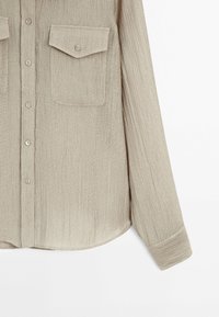Chemise beige à manches longues en tissu texturé, devant boutonné, poches poitrine à rabat boutonné et manches avec poignets.