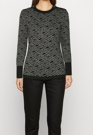 Femme portant un pull à manches longues noir et gris à motifs géométriques, avec des boutons sur l'épaule, assorti à un pantalon noir.