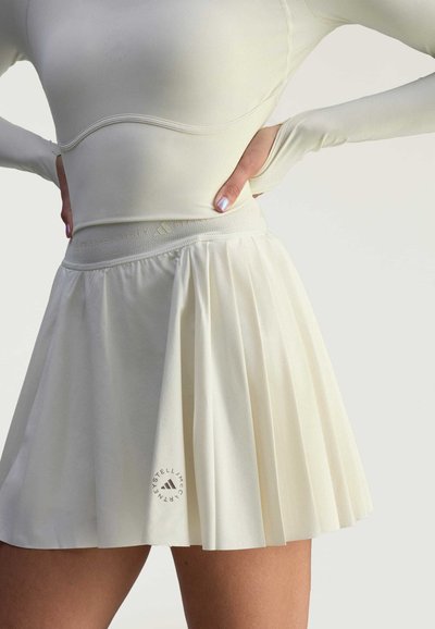 Pleated white skirt with a fitted waistband and textured fabric. Features a small circular logo on the hem, with a smooth, soft appearance.
