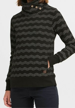 Femme portant un pull noir à rayures blanches en zigzag, col montant boutonné, manches retroussées et poche avant, assorti à un pantalon foncé.