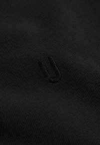 Černá tkanina ukazuje detailní záběr na texturu s jemně vyšitým malým písmem "u". Materiál: hladký, mírně strukturovaný úplet.