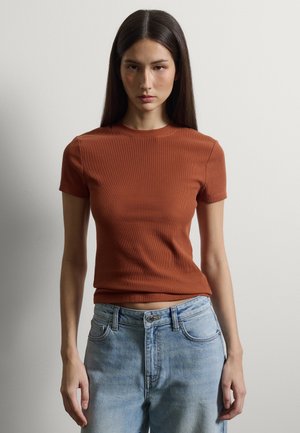 Junge Frau mit langen dunklen Haaren, die ein eng anliegendes, rostfarbenes Kurzarmoberteil und hellblaue hoch taillierte Jeans trägt, vor einer schlichten Wand.