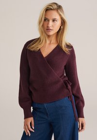 Pull enroulé bordeaux avec un col en V, texture côtelée, manches longues et un accent noué à la taille. Assorti à un pantalon en denim.