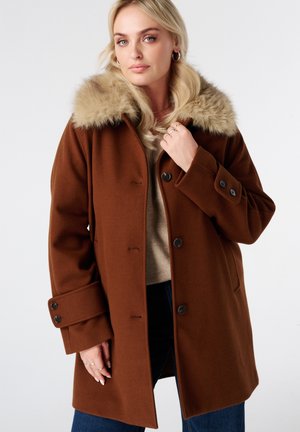 Femme portant un manteau en laine marron avec des boutons noirs et un col en fausse fourrure beige, associé à un jean bleu foncé et un pull beige.