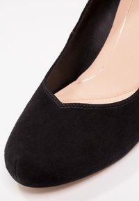 Chaussure à talon haut en daim noir avec un bout pointu et un décolleté bas, présentant des accents cousus le long du bord.