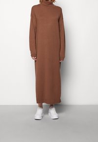 Brun stickad polotröja klänning med långa ärmar, sidofickor och en lös passform, kombinerad med vita sneakers mot en grå bakgrund.