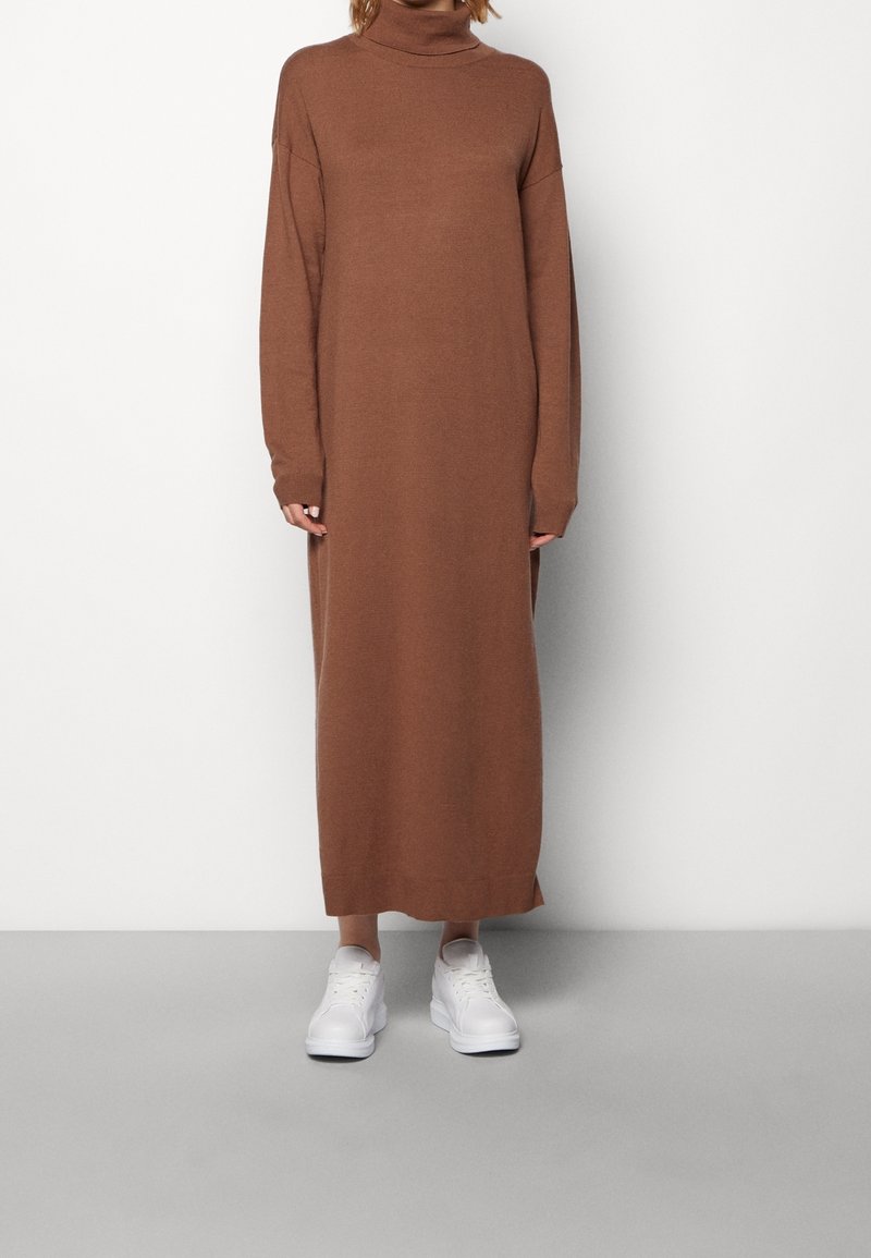 Brun stickad polotröja klänning med långa ärmar, sidofickor och en lös passform, kombinerad med vita sneakers mot en grå bakgrund.