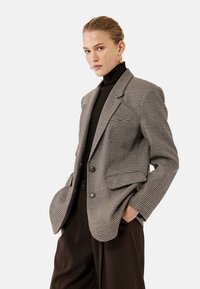 Brunternet blazer med to knapper, spidse klapper og frontlommer, parret med en sort rullekrave. Glat stof med struktureret design.