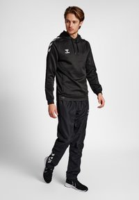 Zwarte hoodie met logo, gecombineerd met zwarte elastische broek met zijkanten in een streep. Het materiaal lijkt soepel en lichtgewicht.