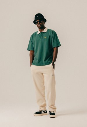 Mann trägt grünes Poloshirt, beige Jogginghose, schwarzen Anglerhut, Sonnenbrille und schwarze Sneakers, steht mit den Händen in den Taschen.