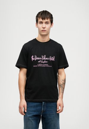 Ung mann med kort, mørkt hår iført en svart T-skjorte med rosa tekst "The Venice Palace Hotel" og blå jeans, som står mot en ensfarget bakgrunn.
