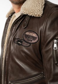Bruine leren jas met een schapenvachtkraag. Voorzien van geborduurde patches en een ritszak, wat een vintage ontwerp tentoonstelt.