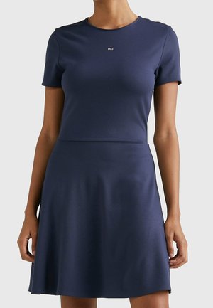 Vrouw draagt een effen marineblauwe A-lijnjurk met korte mouwen en een klein rechthoekig logo op de borst, staand met ontspannen armen.