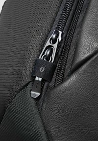 Hoek van een tas van zwart textiel met een structuur en glad leer, met een opvallende metalen rits en een stoffen ritstrekker met een locksymbool.