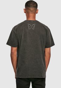 T-shirt en coton gris foncé à manches courtes, avec un design de papillon brodé en blanc sur le haut du dos. Coupe décontractée et col rond.