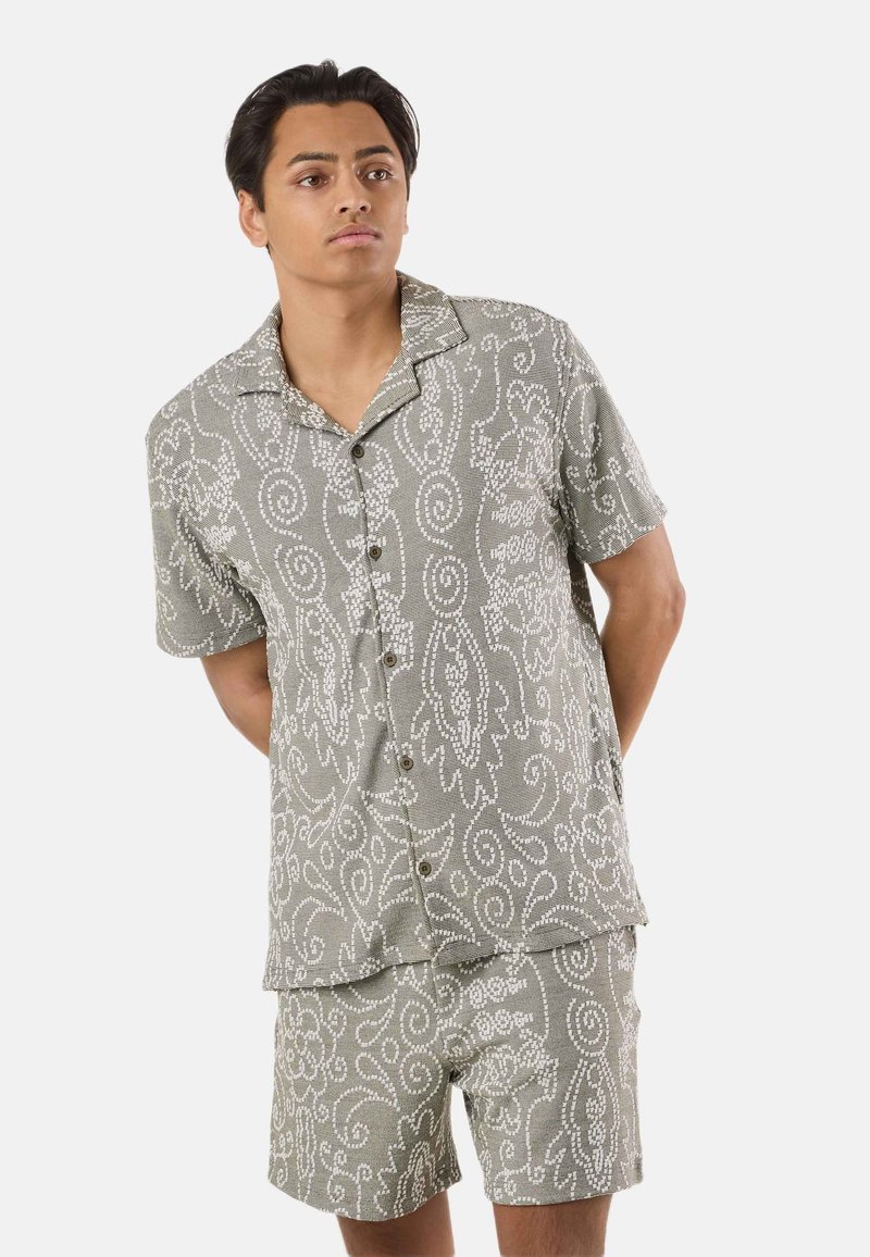 Jeune homme portant une chemise à manches courtes boutonnée et un short assortis, avec des motifs complexes de tourbillons et floraux de couleur claire.