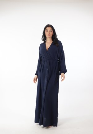 Vrouw met lang donker haar, gekleed in een marineblauwe maxi-jurk met lange mouwen en knopen, staand tegen een witte achtergrond.