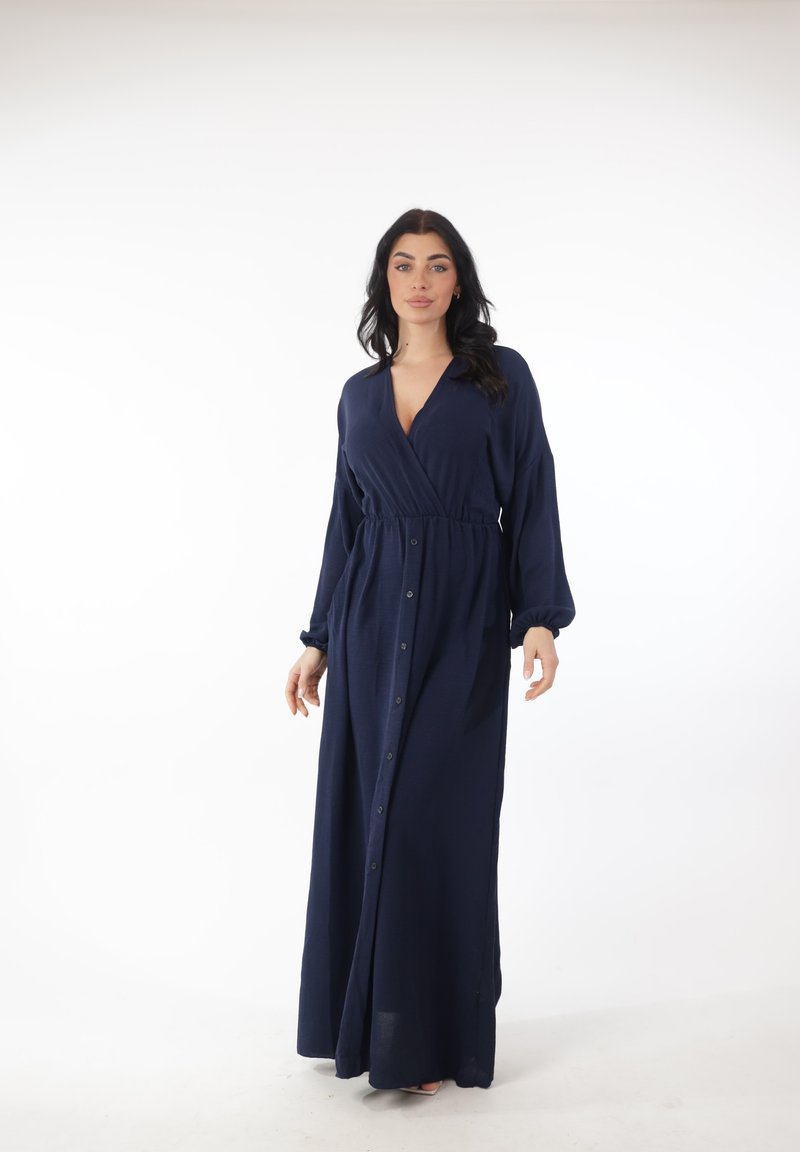 Frau mit langen dunklen Haaren, die ein marineblaues Maxikleid mit langen Ärmeln und Knöpfen trägt, steht vor einem weißen Hintergrund.