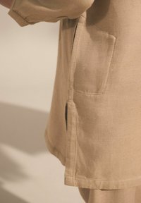 Beżowy strój z boczną kieszenią, wykonany z teksturowanej tkaniny. Brzeg jest wygięty z detalami szwów, charakteryzując się swobodnym stylem.