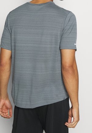 Homme portant un t-shirt gris texturé à manches courtes et un pantalon noir, vu de dos sur un fond clair uni.