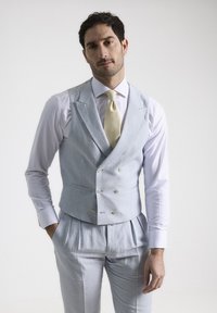 Chaleco de lino azul claro con diseño de doble botonadura, botones plateados y dos bolsillos frontales, combinado con pantalones grises claros y una camisa blanca.