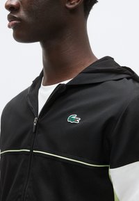 Črna športna jakna na zadrgo s kapuco, ki ima zeleno-belo poudarjeno črto, teksturirano tkanino in majhen vezen krokodilji logotip.