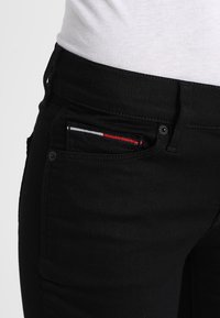 Jeans de mezclilla negros presentan un diseño ajustado con una cintura alta. Los detalles de los bolsillos incluyen una pequeña franja horizontal en rojo, blanco y azul.