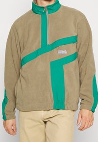 Tan fleece pullover med et grøn geometrisk design, høj krave, halv lynlås og elastiske mancher, med et lille logo på forsiden.