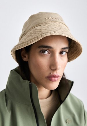 Pălărie bej tip bucket, realizată dintr-un material texturat, cu un logo cusut în față, purtată împreună cu o jachetă verde. Aspectul general este casual și contemporan.