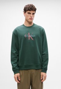 Sweatshirt verde feito de algodão com um colarinho canelado, apresentando um logótipo impresso ao centro que diz "Calvin Klein" em cores contrastantes.