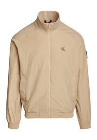 Chaqueta ligera beige con cuello alto, cremallera frontal, puños elásticos y cinturilla; cuenta con un logo en el lado izquierdo del pecho y bolsillos laterales.