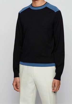 Mann in einem schwarzen Pullover mit blauen Schultern und Saum, kombiniert mit weißen Hosen, vor einem schlichten hellgrauen Hintergrund stehend.