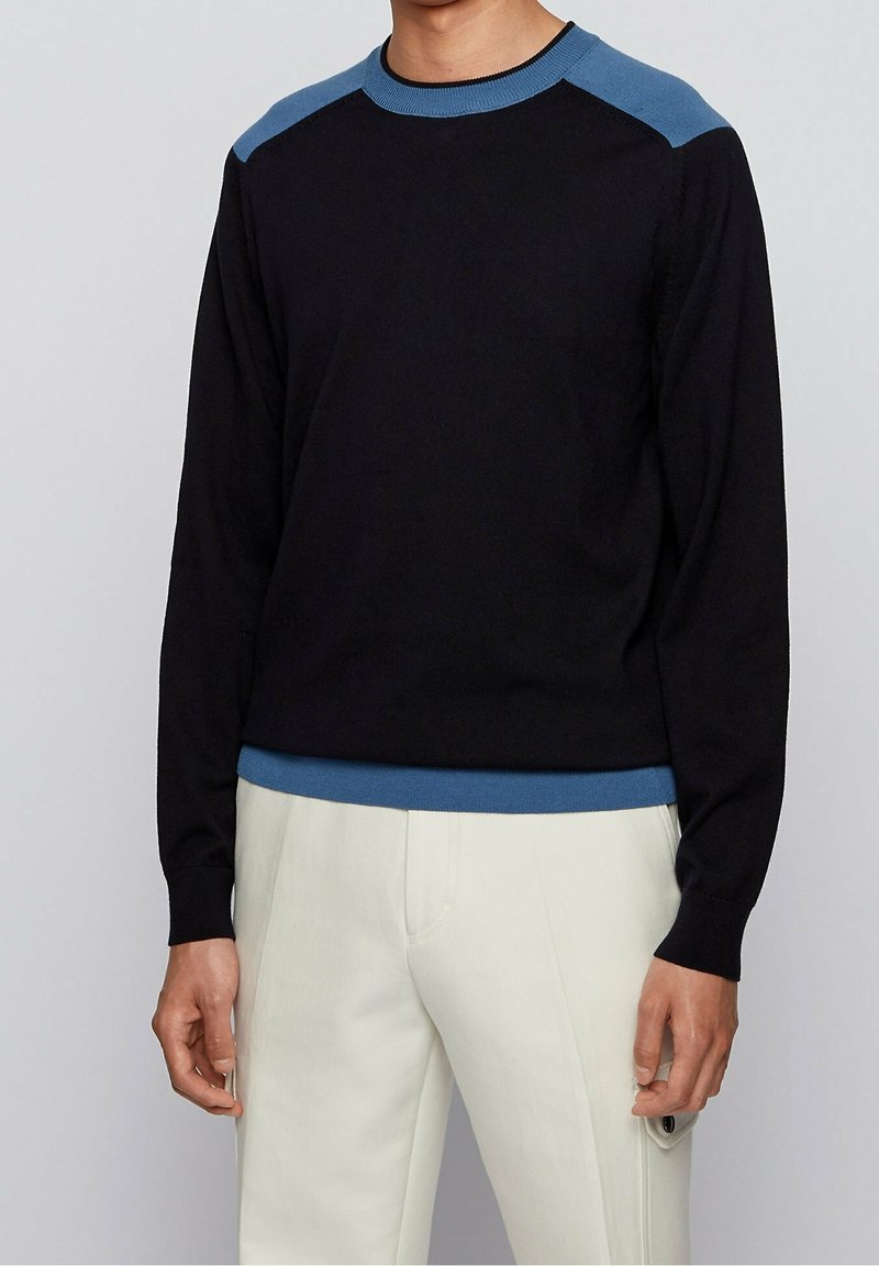 Uomo che indossa un maglione nero con spalle e orlo blu abbinato a pantaloni bianchi, in piedi contro uno sfondo grigio chiaro uniforme.