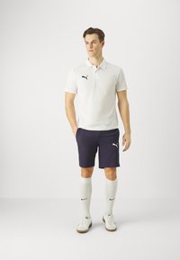 Weißes Polohemd mit schwarzem Puma-Logo, navyfarbene Shorts mit Logo, weiße kniehohe Socken und weiße Sneaker mit beigen Sohlen.