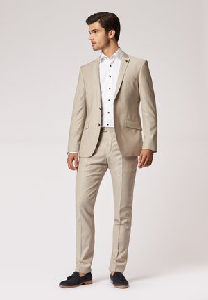 Beige Anzug mit taillierter Passform, bestehend aus einem einreihigen Jackett, mit Revers und einem weißen Hemd mit braunen Knöpfen. Marineblaue Schuhe.