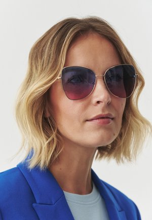 Ochelari de soare tip cat-eye cu lentile degrade de la violet închis la roz deschis, cu rame subțiri aurii, purtați cu un sacou albastru peste un top de un albastru deschis.