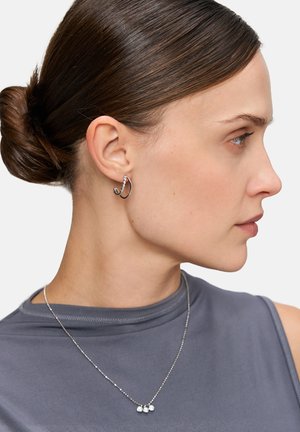 Silberne Creolen mit glitzernden Akzenten, kombiniert mit einer filigranen Kettenkette, die drei kleine, klare Steine aufweist. Graues Stoffoberteil.