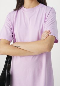 Koszulka z krótkim rękawem i okrągłym dekoltem w jasnopurpurowym kolorze z bawełny, charakteryzująca się luźnym krojem i gładką teksturą, trzymana w torebce w czarnym kolorze.