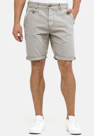 Mand iført lysegrå shorts med opkrogede kanter og knapdetaljer samt hvide sneakers, stående med den ene hånd i lommen.