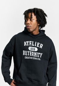 Mustam kootud pehme kangaga kapuutsiga, millel on kenguru tasku ja valge trükitud tekst "ATELIER RETERNITY 2018 LOOVJUHT."