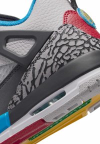Ingrandimento di una sneaker multicolore che mostra pelle grigia e rossa testurizzata, supporto in plastica nera, sovrapposizione stampata e suola bianca con cuscinetto d'aria visibile.