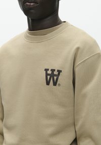Beige sweatshirt lavet af bomuld. Har en rund halsudskæring og et mørkebrun logo på venstre bryst. Blød tekstur med ribbede cuffs.