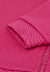 Fuchsiová mikina s ribovanou strukturou, která má ohrnuté manžety a kontrastní zip, s hladkým a příjemným povrchem.