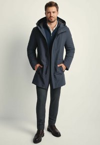 Granatowa parka z zamkiem błyskawicznym z przodu, dużym kapturem i bocznymi kieszeniami; noszona na teksturowanym ciemnym swetrze i eleganckich szarych spodniach. Ciemnobrązowe buty sznurowane.