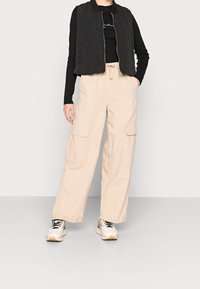 Schwarze gesteppte Weste über einem schwarzen Langarmshirt, kombiniert mit beigen Cargo-Hosen mit Seitentaschen und weißen Sneakers mit schwarzen Akzenten.