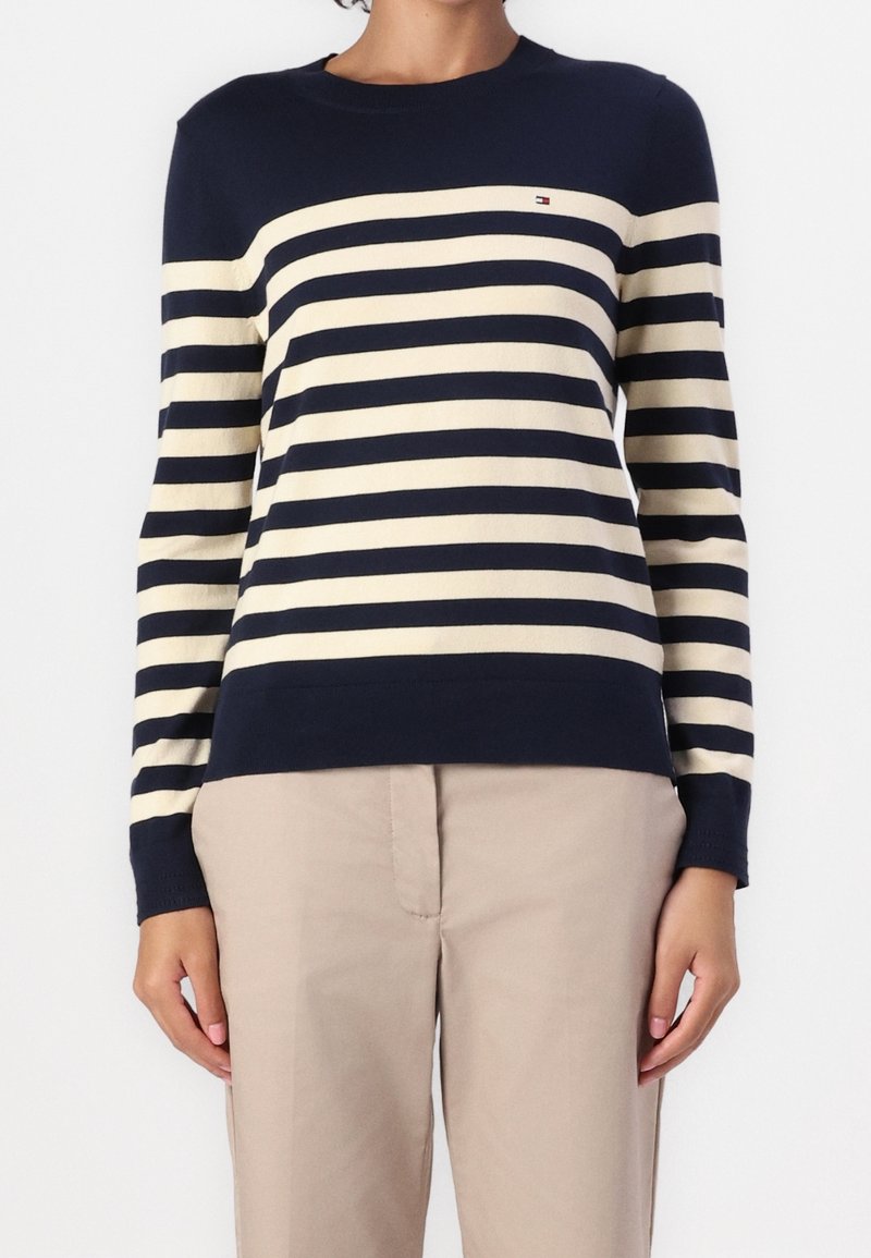 Femme portant un pull à manches longues à rayures horizontales bleu marine et crème, et un pantalon beige, debout devant un fond blanc uni.