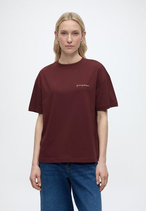 Kobieta o długich blond włosach stoi zwrócona przodem, ubrana w bordową koszulkę z małym białym napisem i niebieskie dżinsy na jednolitym tle.