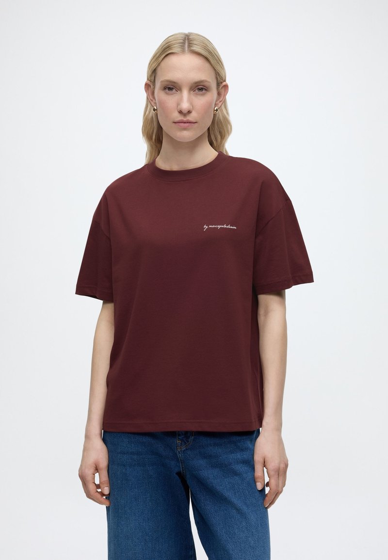 Γυναίκα με μακριά ξανθά μαλλιά στέκεται με το πρόσωπο προς τα εμπρός, φορώντας ένα βυσσινί T-shirt με μικρό λευκό κείμενο και μπλε τζιν, μπροστά σε απλό φόντο.